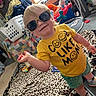 blanket, carpet, casual, child, clothing, floor, happy, indoor, laundry_basket, person, playroom, room, shorts, smiling, sneakers, sunglasses, text_on_shirt, toddler, toys, yellow_shirt