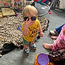 bag, blanket, carpet, child, clothing, colorful, container, feet, floor, furniture, indoor, person, playroom, shoes, smileys, snacks, sunglasses, toddler, toys, yellow_shirt