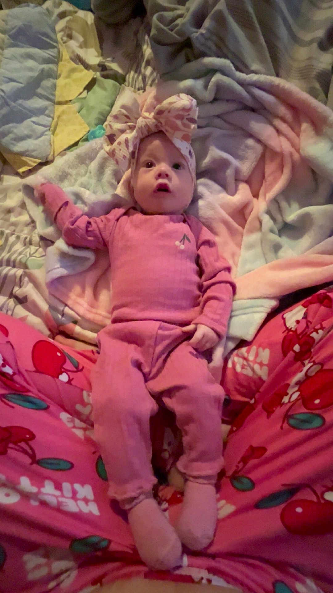 Evelynn joined the competition — help win amazing prizes! baby, infant, newborn, headband, bow, onesie, pink_clothing, socks, blanket, bed, pajamas, cherry_pattern, looking_up, wide_eyes, parent_legs, pacifier, cozy, soft_fabric, portrait, indoor