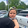 arm, backwards_cap, blurred_background, boy, cap, car, casual, child, clothing, daytime, expression, face, greenery, holding, nature, outdoor, parking_lot, person, photography, vehicle