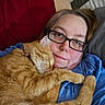 cat, tabby_cat, orange_cat, pet, woman, person, glasses, snuggling, sleeping, relaxing, bed, bedroom, pillows, blanket, blue_shirt, red_pillow, portrait, indoors, close_up, cuddling