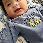 Jin is registered to the contest to win money with this photo: baby, smiling, infant, face, hand, blanket, clothing, shirt, text, indoor, cozy, cute, young_child, happy, portrait, lying_down, soft_texture, patterned_blanket, demolition_expert, black_hair