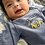 baby, smiling, infant, face, hand, blanket, clothing, shirt, text, indoor, cozy, cute, young_child, happy, portrait, lying_down, soft_texture, patterned_blanket, demolition_expert, black_hair