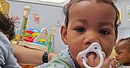 Roy is registered to the contest to win money with this photo: toddler, child, pacifier, indoor, daycare, toys, bookshelf, colorful, playroom, infant, baby, person, furniture, window, wall_decor, sitting, young_child, cute, hand, expression