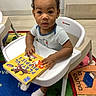 Roy is registered to the contest to win money with this photo: baby, child, toddler, high_chair, book, reading, indoor, carpet, colorful, playful, curious, person, clothing, socks, floor, toy, education, early_learning, cute, portrait