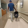 man, baby, newborn, car_seat, hospital, hallway, walking, blue_shirt, white_pants, cap, tattoo, floor, walls, picture_frame, clock, door, light, person, infant, medical