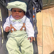 Jeremiah is registered to the contest to win money with this photo: baby, stroller, child, hat, bow_tie, suspenders, pants, shirt, footwear, floor, wooden_floor, indoor, person, face, cute, seated, clothing, hand, expression, black_skin