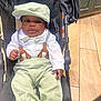 baby, stroller, child, hat, bow_tie, suspenders, pants, shirt, footwear, floor, wooden_floor, indoor, person, face, cute, seated, clothing, hand, expression, black_skin