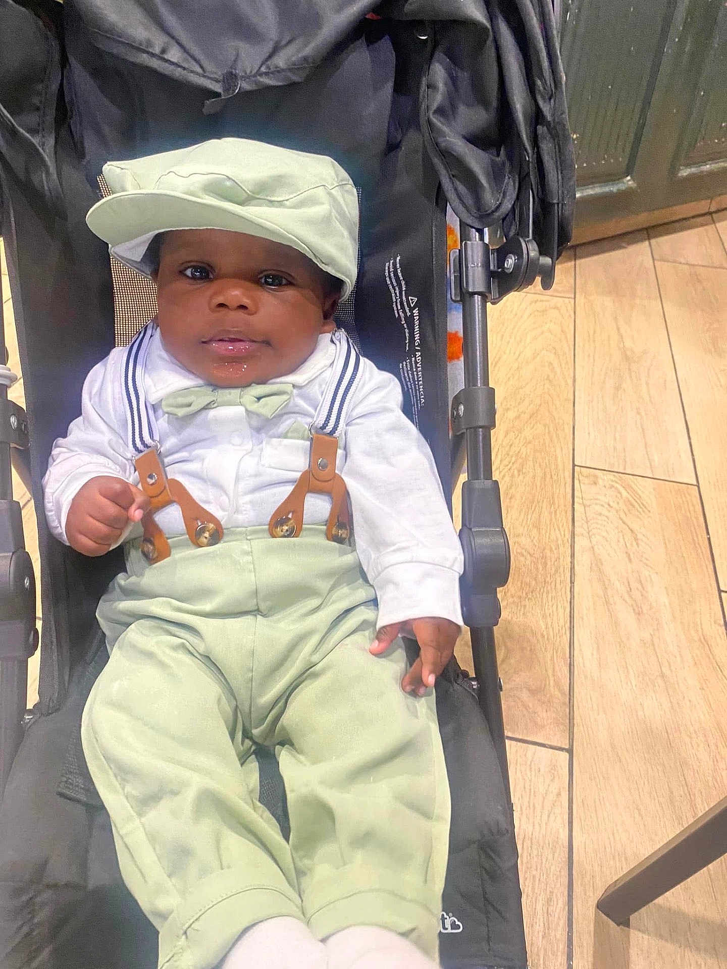 Jeremiah is registered to the contest to win money with this photo: baby, stroller, child, hat, bow_tie, suspenders, pants, shirt, footwear, floor, wooden_floor, indoor, person, face, cute, seated, clothing, hand, expression, black_skin