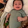 Mateo is registered to the contest to win money with this photo: baby, blanket, child, comfort, cozy, cute, expression, face, green_onesie, hair, happy, indoors, infant, lying_down, person, pillow, skin, smiling, soft, toddler
