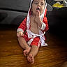 baby, boxing_robe, child, couch, curious, cute, expression, finger_in_mouth, floor, home, indoors, living_room, person, red_clothing, sitting, toddler, toy, white_trim, wooden_floor, young_child