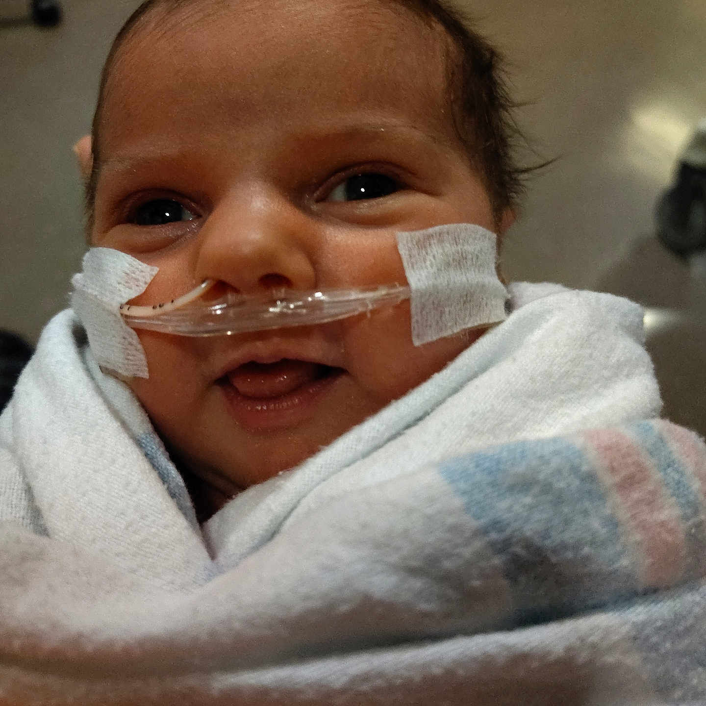 Hazel is registered to the contest to win money with this photo: baby, infant, smile, face, blanket, hospital, nasal_cannula, medical_tape, healthcare, wrapped, newborn, indoors, closeup, cute, person, child, soft_texture, portrait, happy, lying_down