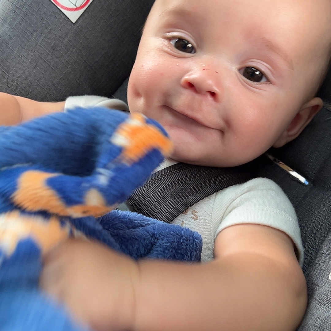 Tatyum is registered to the contest to win money with this photo: baby, baby_clothing, blanket, blue_blanket, car_seat, child, close_up, cute, dimple, face, happy, human, indoors, infant, orange_pattern, portrait, seatbelt, skin, smiling, soft_toy