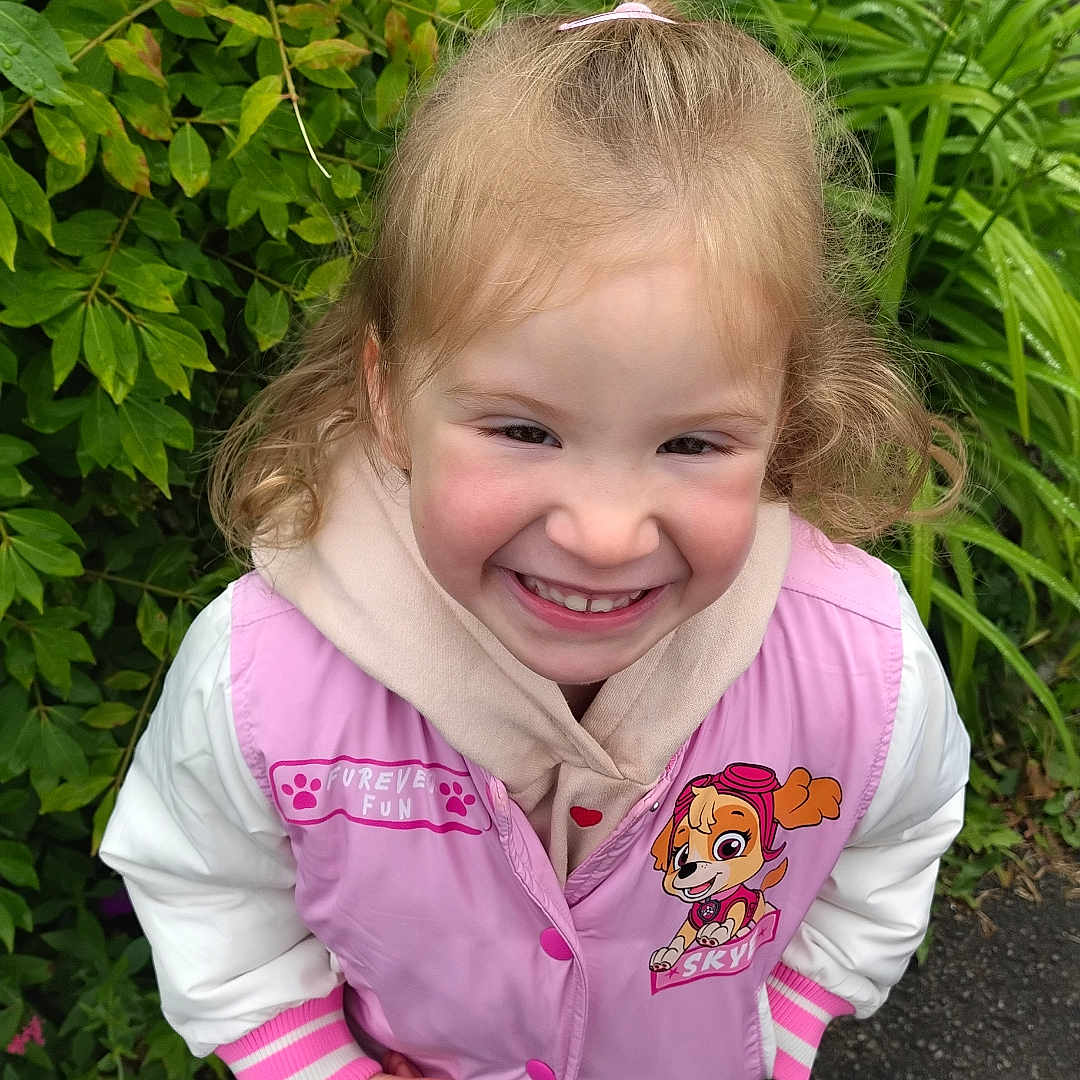 Ambre a rejoint le concours — aidez-le/la à gagner de superbes lots ! casual_clothing, child, curly_hair, cute, daylight, girl, greenery, happy, hoodie, jacket, nature, outdoor, person, pink, plants, playful, portrait, smiling, sneakers, standing