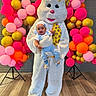 baby, child, easter_bunny, mascot, balloons, balloon_arch, indoor, costume, celebration, party, festive, floor, brick_wall, person, white_costume, yellow_bow, blue_clothing, smiling, headwear, holding