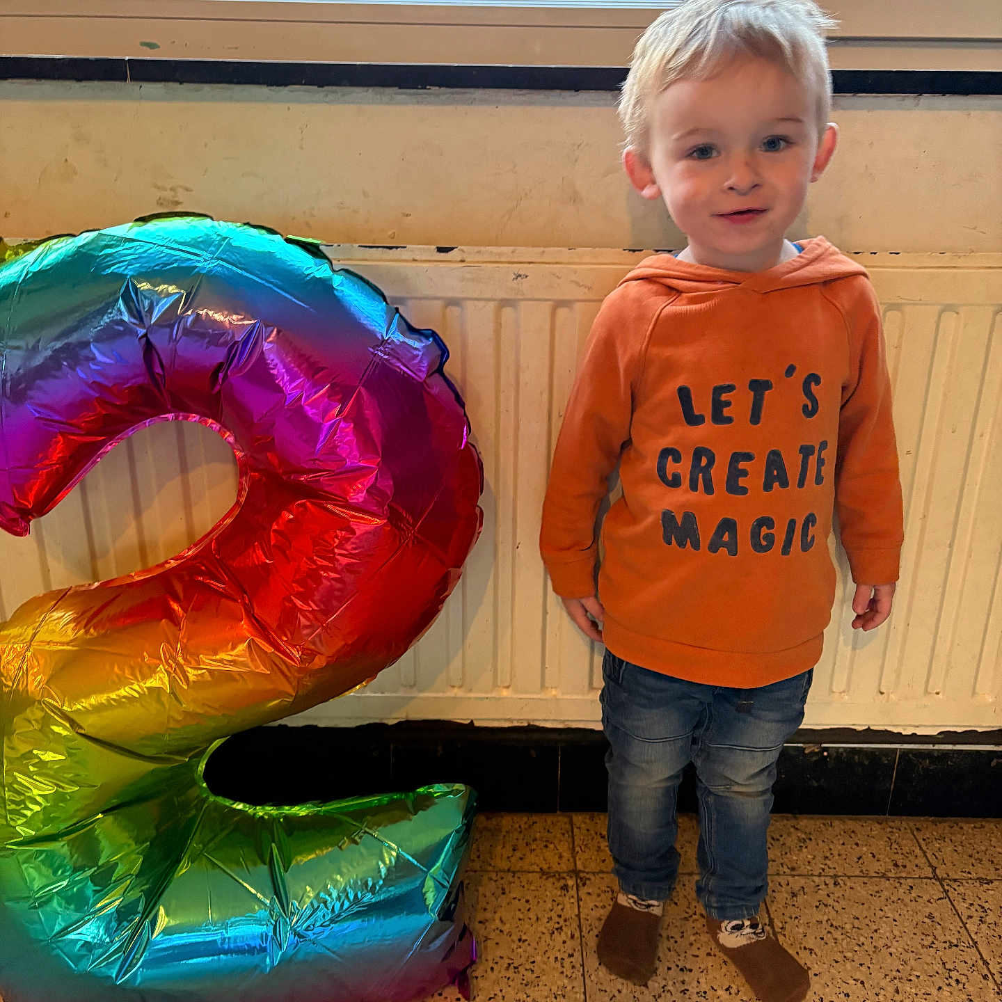 Athanael a rejoint le concours — aidez-le/la à gagner de superbes lots ! balloon, birthday, casual_clothing, celebration, child, cute, floor, happy, hoodie, indoor, jeans, number_two, person, radiator, rainbow, smiling, socks, standing, toddler, window