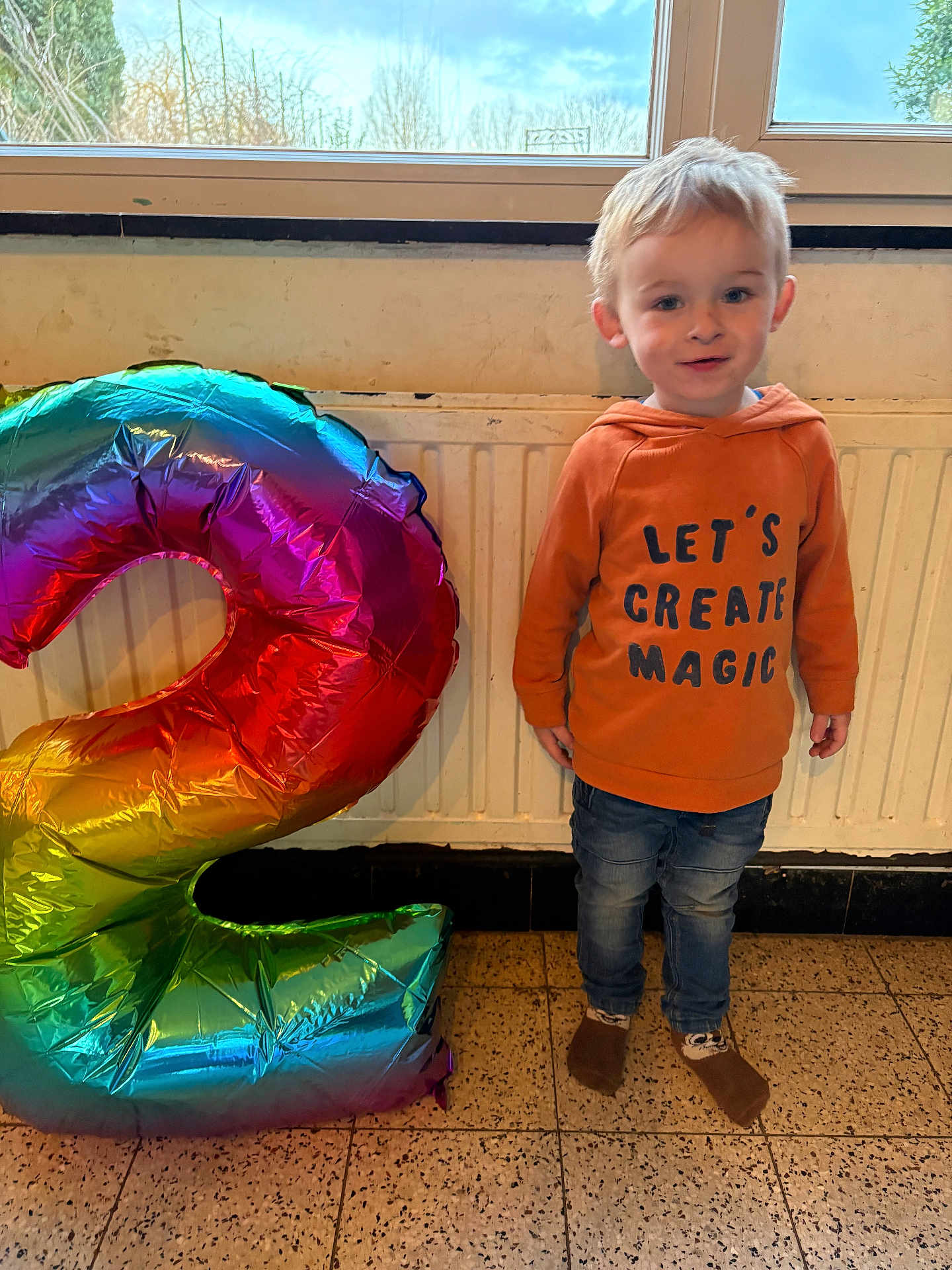 Athanael a rejoint le concours — aidez-le/la à gagner de superbes lots ! toddler, child, balloon, number_two, rainbow, indoor, hoodie, jeans, socks, smiling, floor, window, radiator, celebration, birthday, standing, casual_clothing, happy, person, cute