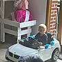 baby, blue_flowers, brick_wall, child, childhood, decor, floor_tiles, indoor, mannequin, natural_light, pink_dress, play, seat, serious_face, steering_wheel, toddler, toy_car, white_car, window, young_child