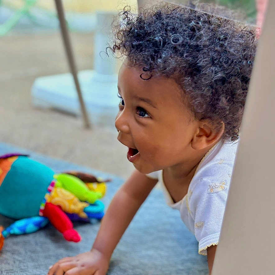 Jahlone participe au concours pour gagner de l'argent avec cette photo : background, blue_rug, child, clothing, colorful, crawling, curly_hair, expression, fabric, face, hand, happy, outdoor, pattern, person, playful, smiling, toddler, toy, young_child