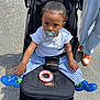 baby, black_stroller, blue_shoes, casual_clothing, child, curly_hair, expression, feet, legs, outdoor, pacifier, pavement, person, striped_pants, stroller, sunlight, toddler, toy_ring, white_shirt, young_child
