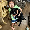 child, girl, cat, black_cat, pet, indoors, living_room, wood_floor, furniture, chair, table, couch, holding, portrait, smiling, long_hair, green_shirt, shorts, skeleton, family_home