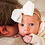 baby, infant, headband, bow, blue_eyes, newborn, close_up, portrait, holding, shoulder, adult, ear, hair, skin, tiny_hand, clothing, cozy, soft_light, indoors, curious_expression