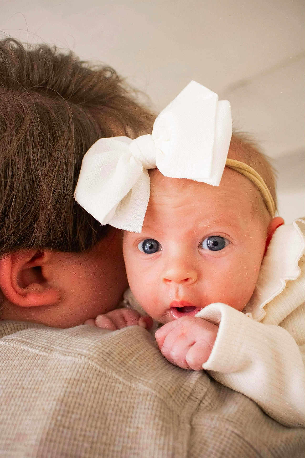 Athena joined the competition — help win amazing prizes! baby, infant, headband, bow, blue_eyes, newborn, close_up, portrait, holding, shoulder, adult, ear, hair, skin, tiny_hand, clothing, cozy, soft_light, indoors, curious_expression