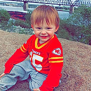 Chase joined the competition — help win amazing prizes! child, smiling, red_jersey, sitting, sand, outdoor, river, bridge, grass, trees, sky, cloudy, happy, young_child, casual_clothing, nature, daytime, portrait, cute, sports_jersey
