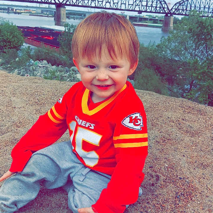 Chase joined the competition — help win amazing prizes! child, smiling, red_jersey, sitting, sand, outdoor, river, bridge, grass, trees, sky, cloudy, happy, young_child, casual_clothing, nature, daytime, portrait, cute, sports_jersey