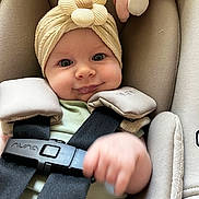Chloe is registered to the contest to win money with this photo: baby, child, car_seat, headband, flower, smile, infant, seatbelt, cute, hand, face, clothing, safety, young_child, indoors, soft_lighting, close_up, happy, person, seat