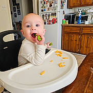 Seth is registered to the contest to win money with this photo: baby, high_chair, kitchen, vegetable, eating, child, face, mouth_open, table, food, refrigerator, cabinets, magnet, photo, person, indoor, seat, clothing, expression, curious