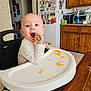 baby, high_chair, kitchen, vegetable, eating, child, face, mouth_open, table, food, refrigerator, cabinets, magnet, photo, person, indoor, seat, clothing, expression, curious