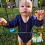 baby, child, onesie, playpen, fence, outdoor, grass, toy, curious, standing, yard, metal, blue_clothing, infant, hands, face, expression, daylight, nature, background