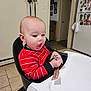 Seth is registered to the contest to win money with this photo: baby, child, high_chair, red_sweater, toy, kitchen, floor_tiles, fridge, magnet, doorway, wall, light_switch, curious, expression, indoor, person, furniture, home, seated, holding