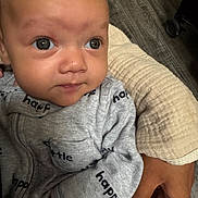 Micah is registered to the contest to win money with this photo: baby, infant, close_up, face, eyes, onesie, clothing, hand, adult_hand, floor, wood_floor, cushion, gray_clothing, text_on_clothing, indoor, portrait, person, cute, young_child, holding
