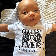 Micah is registered to the contest to win money with this photo: baby, infant, onesie, white_clothing, blanket, lying_down, face, cute, child, soft_texture, home, newborn, portrait, smiling, arms_outstretched, skin, person, sleepwear, toddler, closeup
