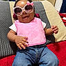 accessory, baby, casual_clothing, child, cute, face, glasses, happy, indoor, infant, jeans, person, pillow, pink_bib, playful, portrait, red_blanket, seated, smiling, sunglasses