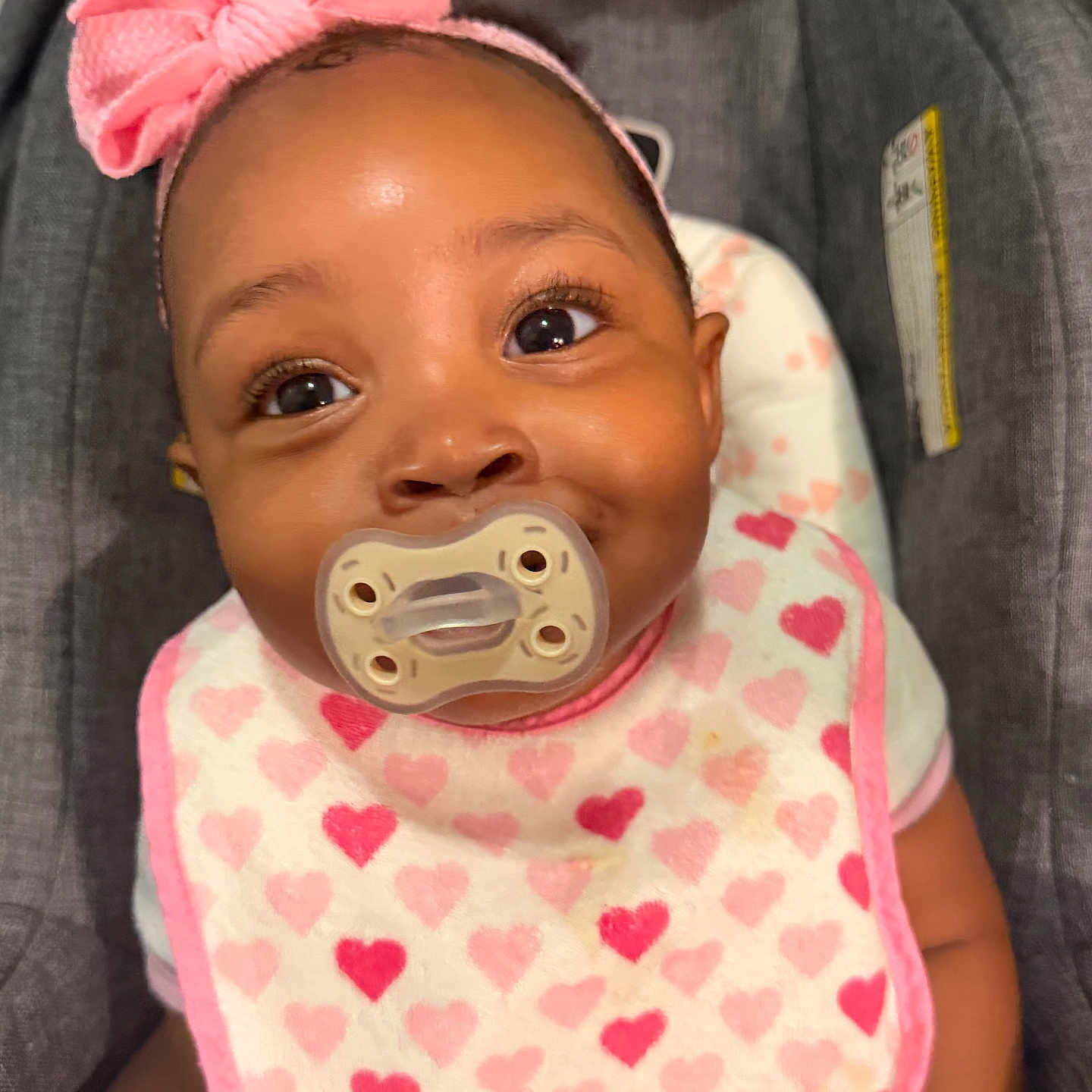 Najiah joined the competition — help win amazing prizes! adorable, baby, baby_accessory, bib, car_seat, child, closeup, clothing, cute, happy, headband, heart_pattern, indoor, infant, pacifier, person, pink_bow, portrait, seat, smiling_eyes