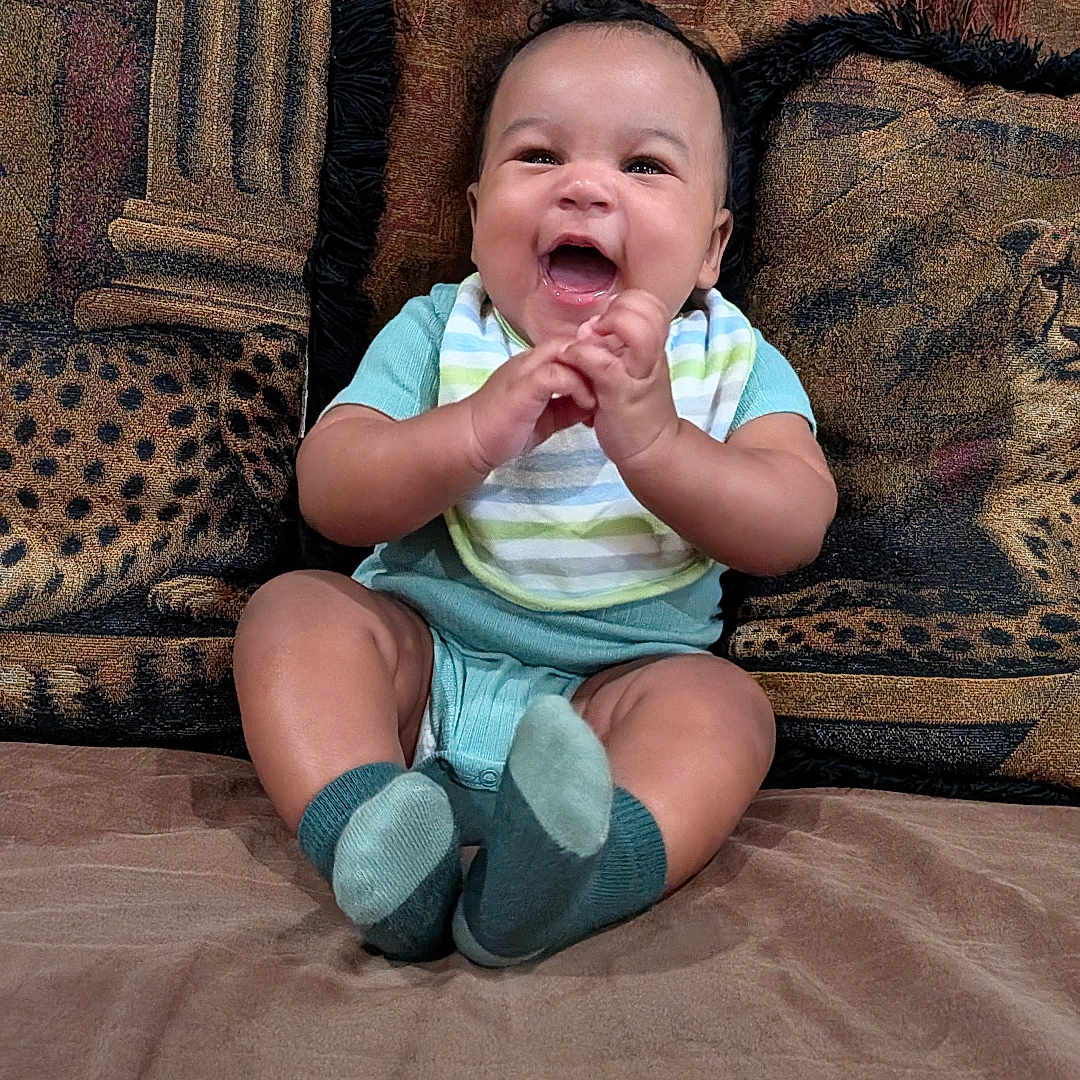 Akeem is registered to the contest to win money with this photo: baby, child, infant, smiling, laughing, sitting, portrait, indoors, couch, cushions, brick_wall, clothing, striped_shirt, shorts, socks, hands, happy, home, cozy, photography