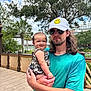 baby, man, child, holding, smiling, hat, glasses, outdoor, trees, wooden_deck, greenery, person, casual_clothing, smiley_face, sky, happy, nature, portrait, daylight, family