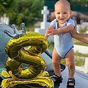 Iavai'Atea participe au concours pour gagner de l'argent avec cette photo : baby, child, balloon, number_8, golden_balloon, crown_balloon, car, hood, hand, support, outdoor, greenery, cross, onesie, sandals, reflection, smiling, celebration, sunlight, portrait