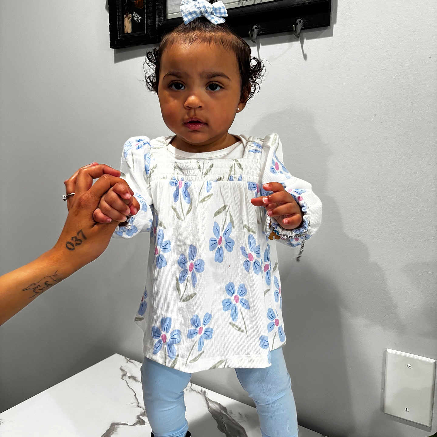 Zoralis Lynn is registered to the contest to win money with this photo: blue_leggings, child, curly_hair, family_happiness, floral_top, hair_bow, hand, holding_hand, indoor, key_decoration, light, marble_surface, person, portrait, standing, table, tattoo, toddler, wall_art, white_shoes