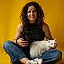 woman, portrait, curly_hair, smile, black_cat, white_cat, cat, pets, pet_owner, sitting, cross_legged, yellow_background, studio, jeans, sneakers, hands, lap, indoor, happy, animal