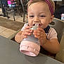toddler, baby, bottle, headband, high_chair, indoor, restaurant, table, person, child, smiling, happy, food, drink, furniture, seat, clothing, people, background, casual