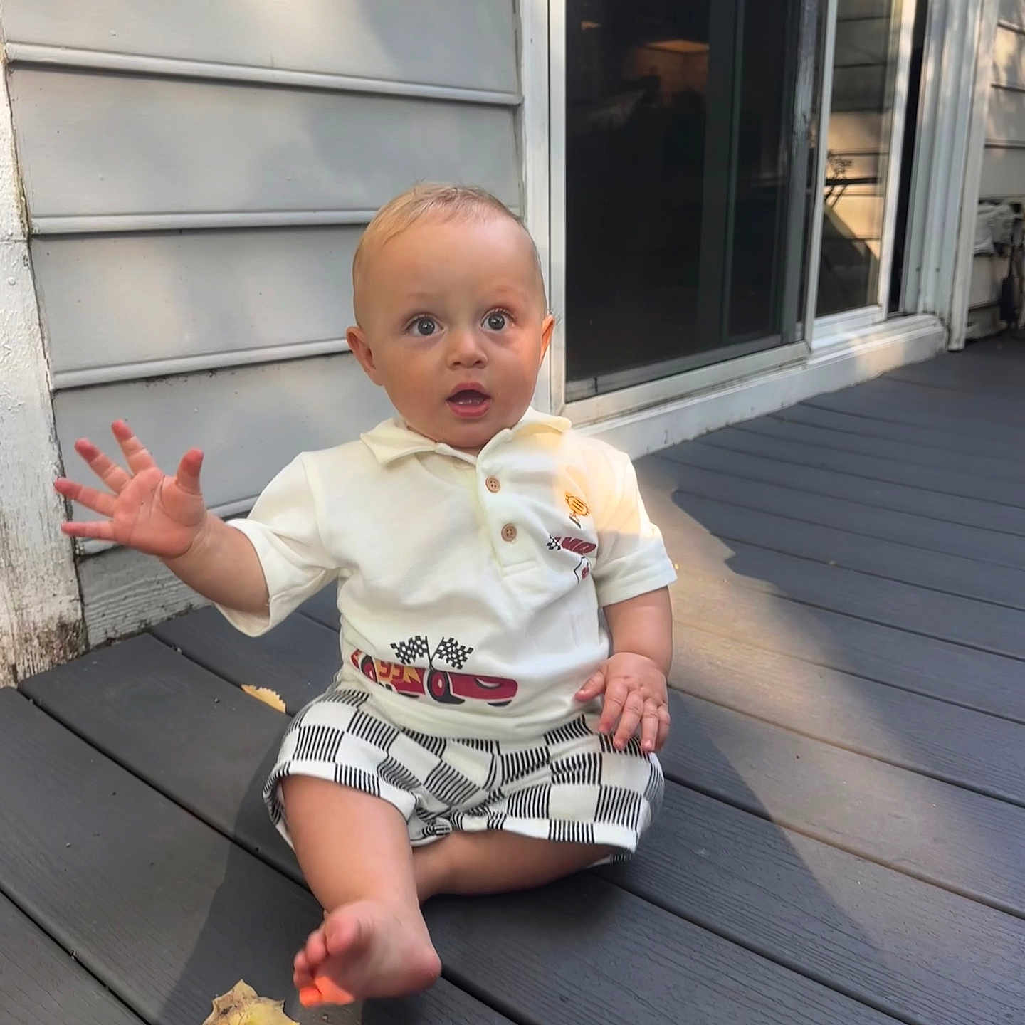 Sam is registered to the contest to win money with this photo: baby, child, outdoor, deck, wood, shirt, shorts, waving, expression, sunlight, shadow, house, window, door, plank, sitting, person, cute, summer, casual
