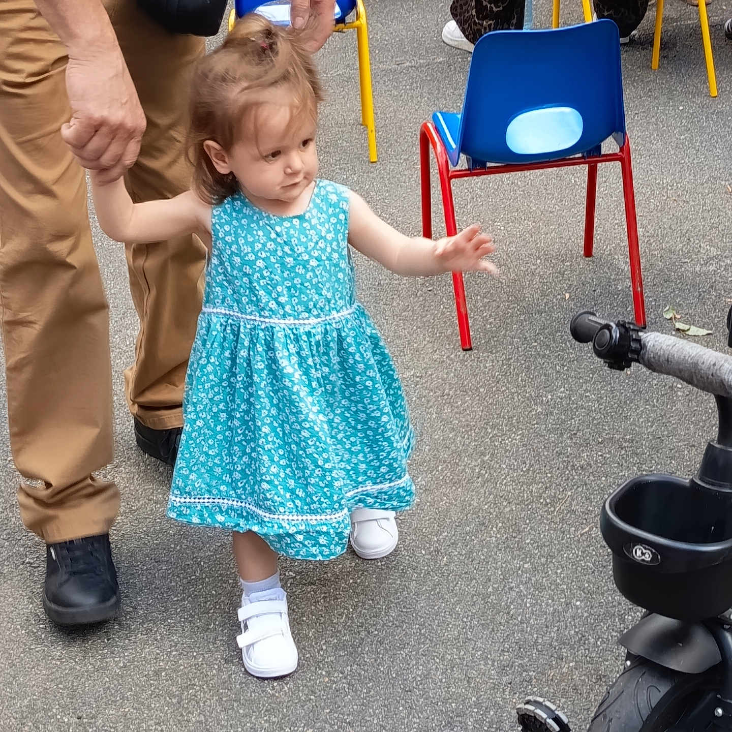 Mia participe au concours pour gagner de l'argent avec cette photo : adult_hand, blue_dress, chairs, child, colorful_chairs, dress, footwear, human_hand, learning_to_walk, outdoor, pavement, person, small_vehicle, support, toddler, toy, tricycle, walking, white_shoes, young_child