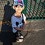 black_purse, cap, casual, child, fashion, frozen, green_fence, mickey_mouse, outdoor, pavement, person, shadow, smiling, standing, summer_clothing, sunglasses, sunlight, tshirt, white_sandals, young_child