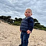 toddler, child, beach, sand, cloudy_sky, trees, outdoor, puffer_vest, jeans, sneakers, nike, casual_clothing, young_child, nature, landscape, standing, portrait, daytime, overcast, person