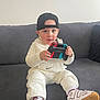 toddler, child, sitting, couch, controller, video_game, cap, white_clothing, sneakers, indoor, person, gaming, casual, footwear, hoodie, cute, focused, modern, living_room, playful
