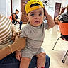 baby, child, infant, portrait, sitting, yellow_cap, gray_outfit, sneakers, wide_eyes, indoors, waiting_room, adult, orange_chair, tile_floor, doors, wall_fan, family, fashion, close_up, photography